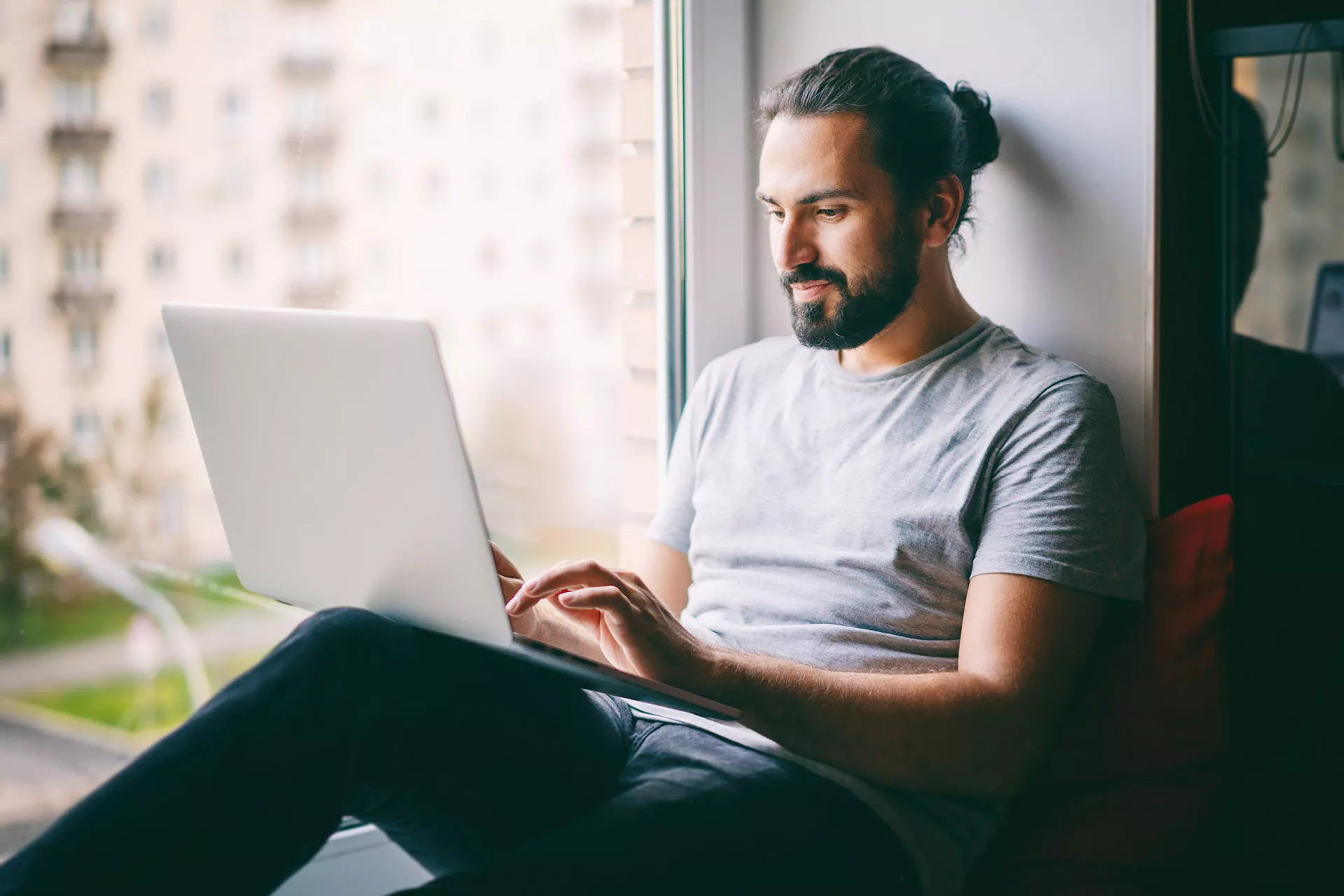 Junger Mann sitzt am Fenster mit Aussicht auf Stadt und arbeitet auf Laptop